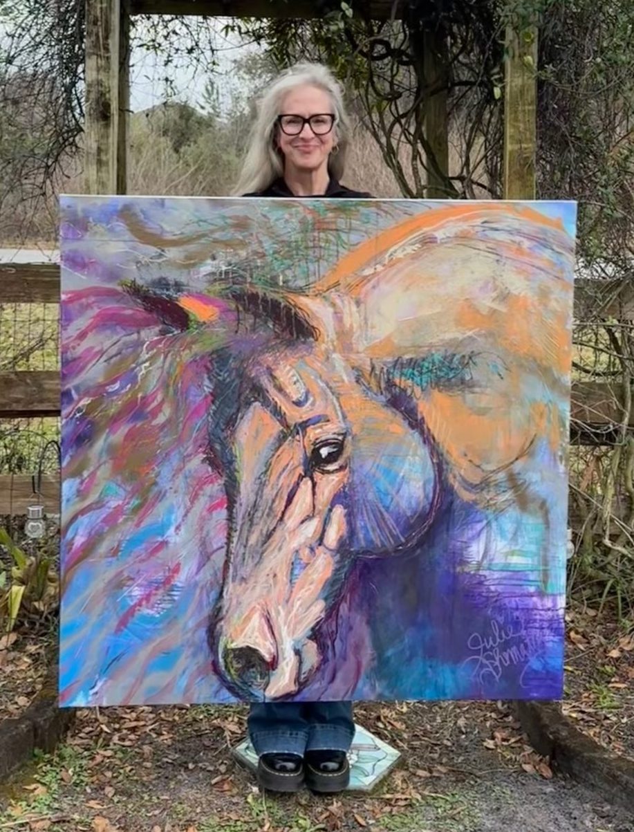 Photo of Julie with one of her paintings in front of a gate in her yard